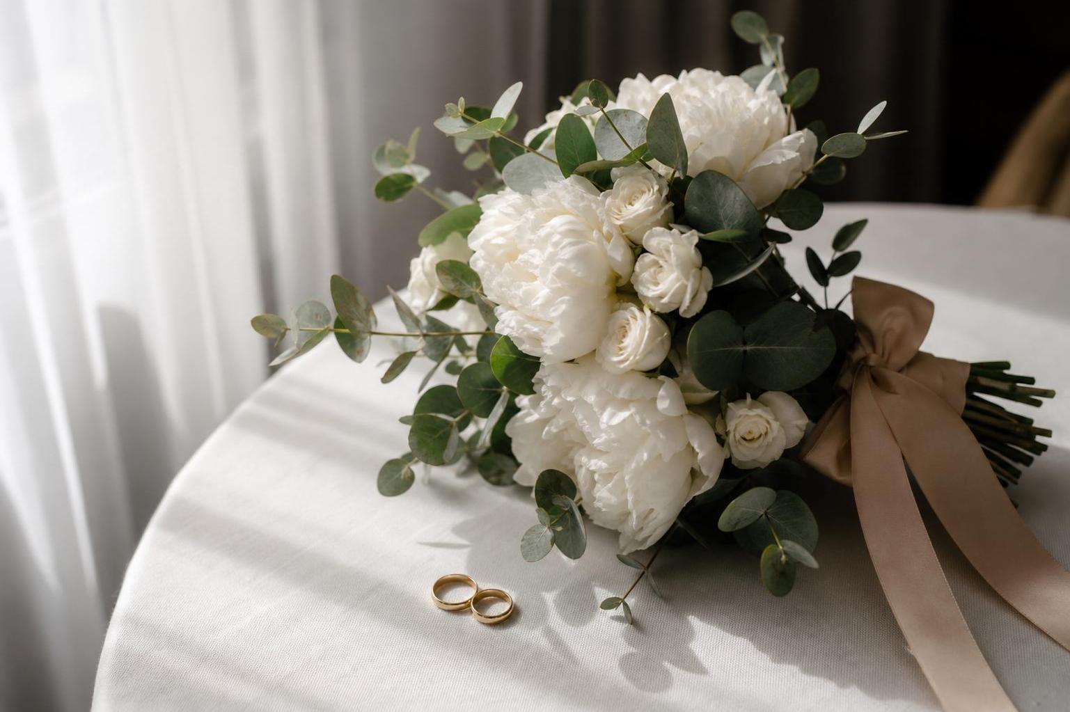 a bridal bouquet of white peonies, White O'Hara spray roses and silver eucalyptus with nude silk ribbon lying on a white tablecloth next to wedding rings, soft morning light