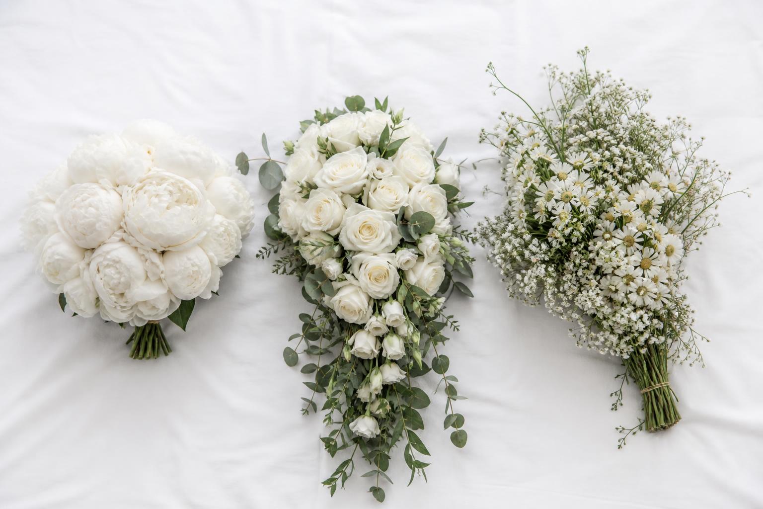 three bridal bouquets of different shapes side by side on white fabric — round dome of peonies, cascade of white roses and greenery, asymmetric 'wildflower' style with gypsophila, soft morning light