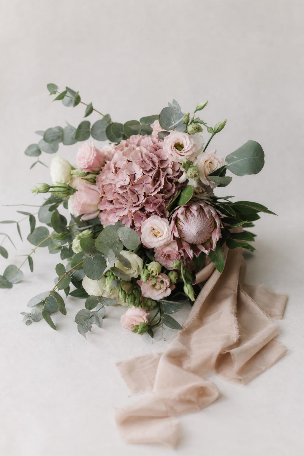 an asymmetric bridal bouquet of hydrangeas, lisianthus, eucalyptus and protea with a long trailing silk ribbon on a white studio background
