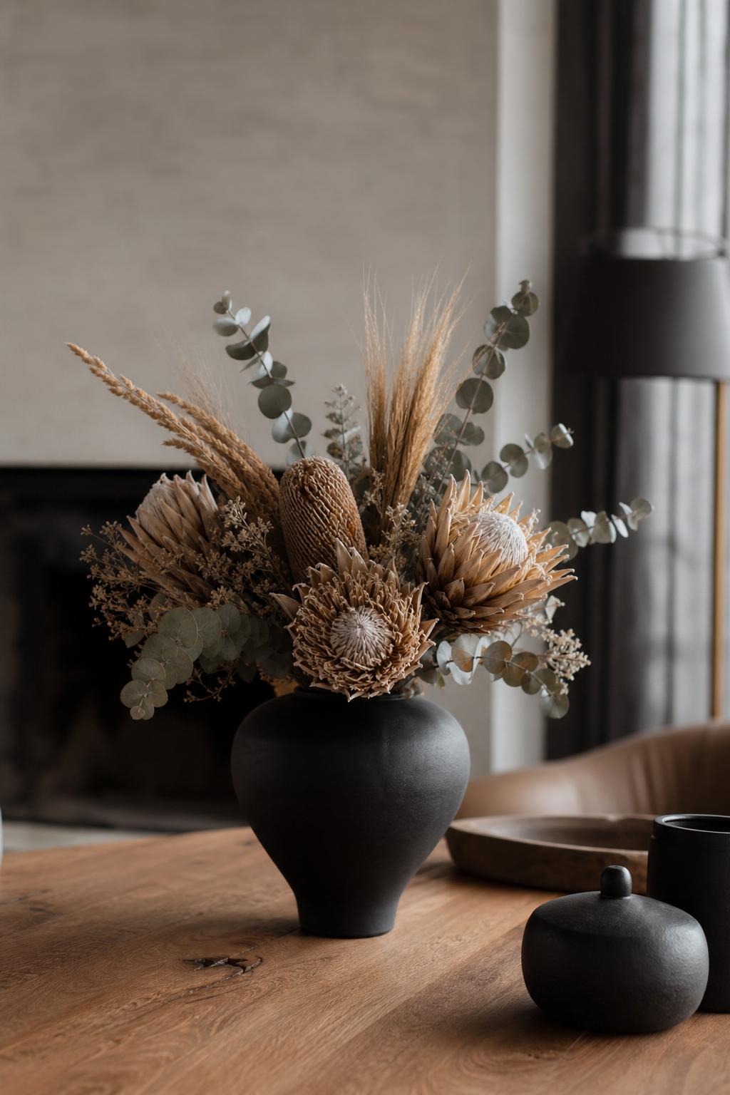 a dried composition of proteas, Banksia, dried wheat, and Baby Blue eucalyptus in a matte black ceramic vase, minimalist designer interior