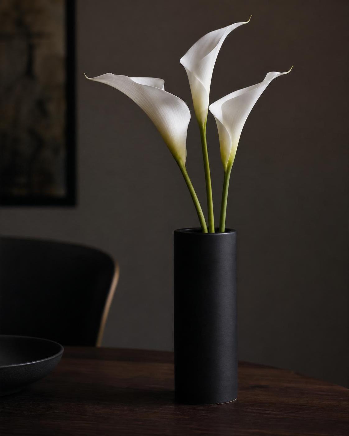 three white calla lilies in a tall matte black ceramic vase on a dark wooden table, minimalist architectural still-life