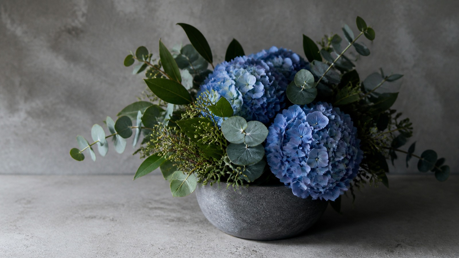 a compact arrangement of blue hydrangea with eucalyptus and dark green foliage in a grey stoneware vessel on a concrete surface, cool directional light