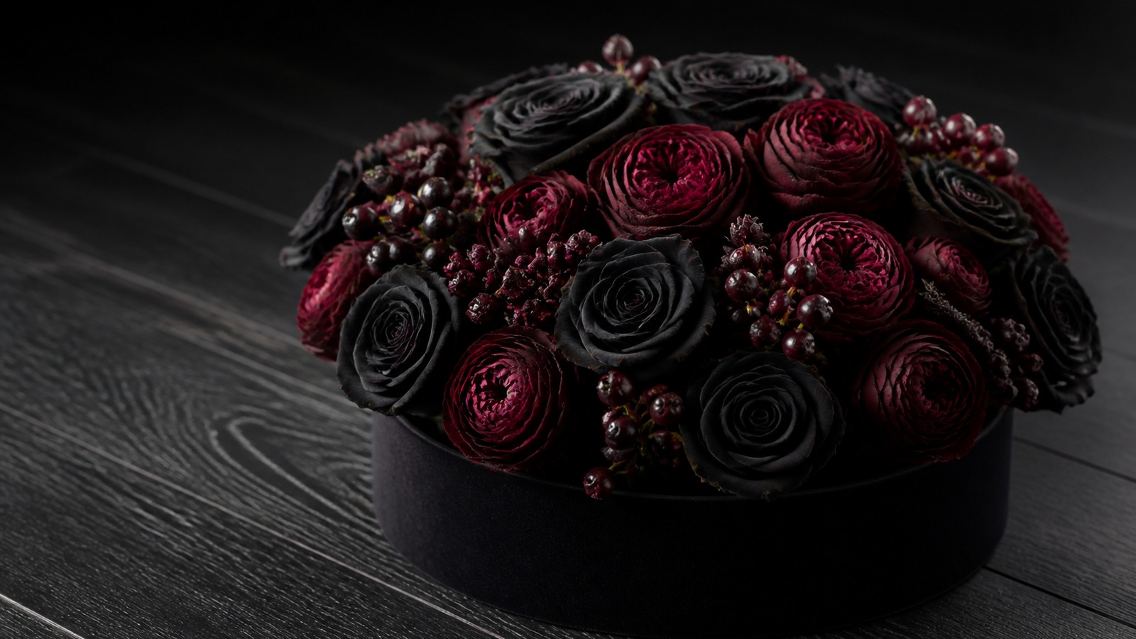 a hatbox arrangement of Black Baccara roses and burgundy ranunculus with dark hypericum berries on a matte black surface, moody spotlight, luxurious editorial