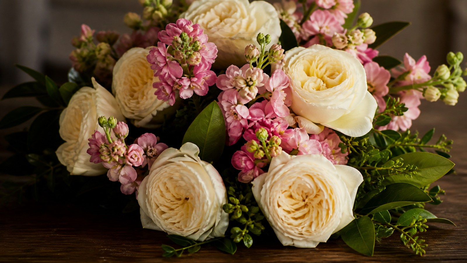 a low compact bouquet of cream garden roses and pink stock on a dark wooden table, deep green foliage, warm soft light, close crop showing petal texture