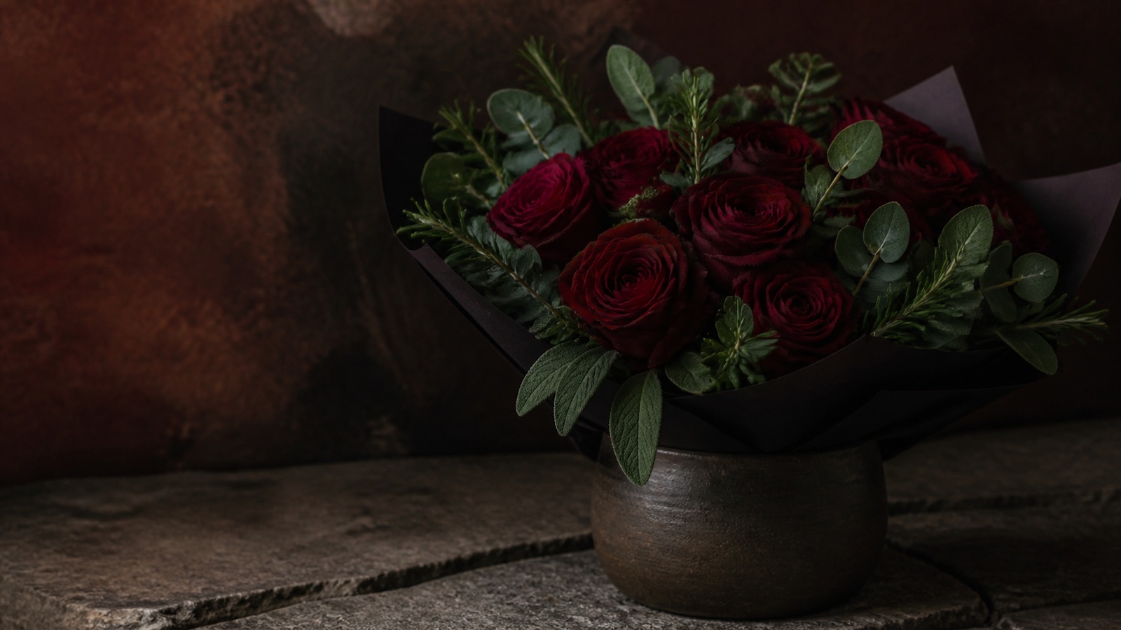a compact bouquet of deep red roses with rosemary, sage and eucalyptus in dark matte paper, standing in a heavy stoneware vessel on a stone surface, low directional light